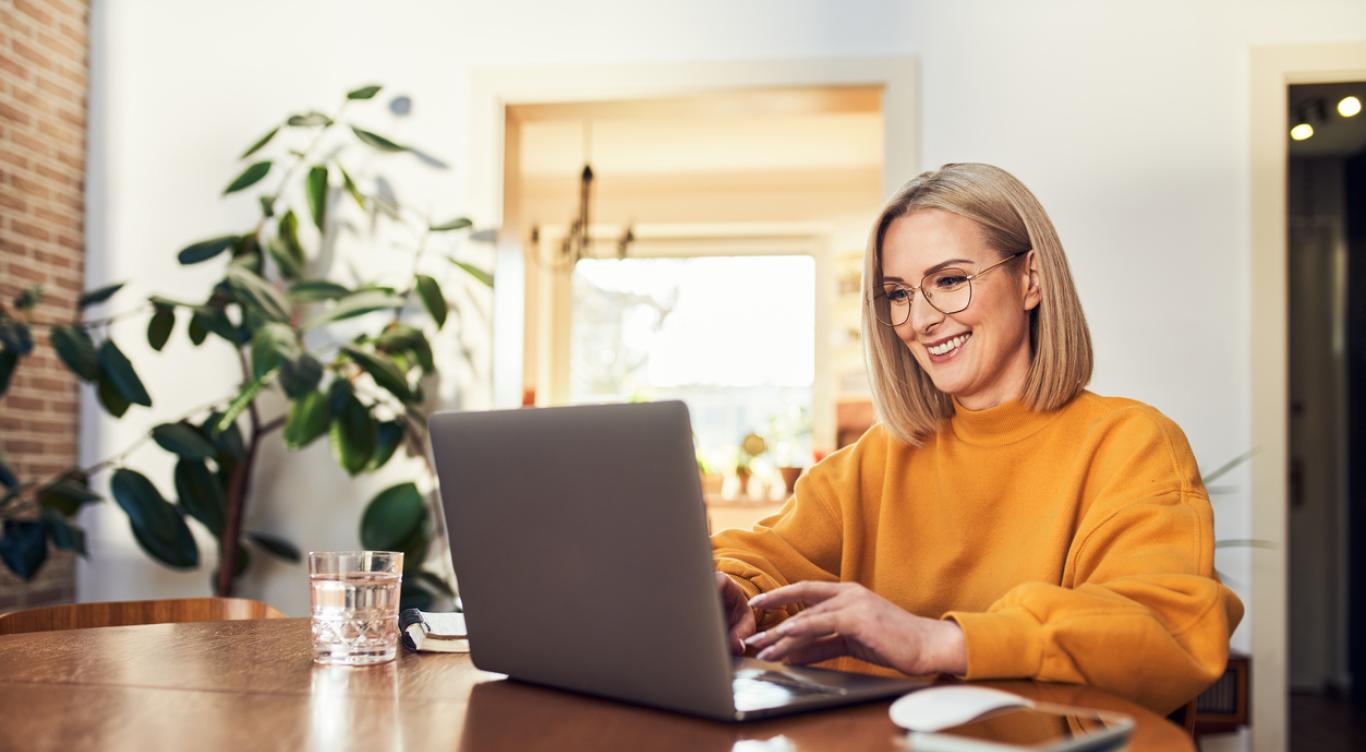 Woman sits at table smiling while looking at computer